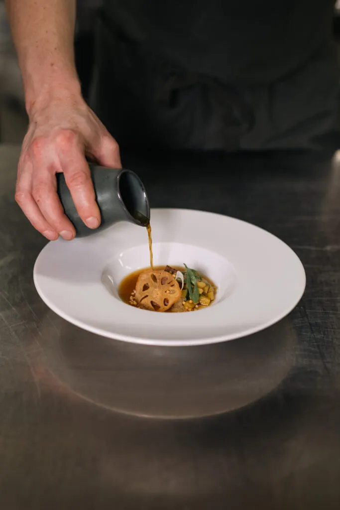 Kock häller mörk sås från en liten karaff över en elegant tallrik med lotus root och grönsaker på vit porslinstallrik