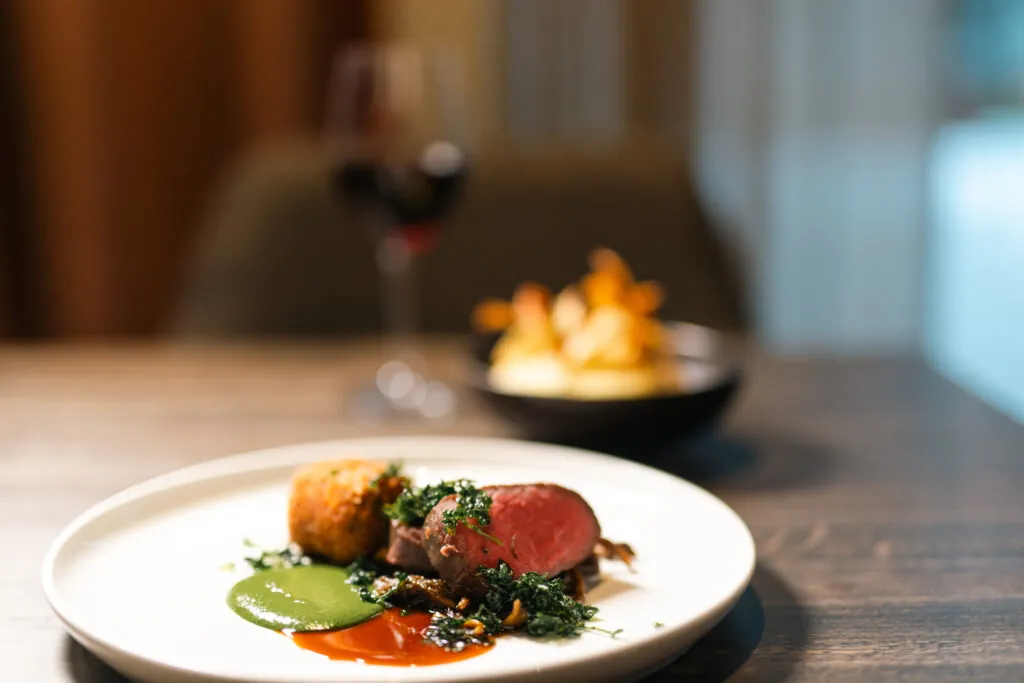 Elegant kött med grön sås, kroketter och krispig sallad på vit tallrik, serverat i restaurangens varma atmosfär
