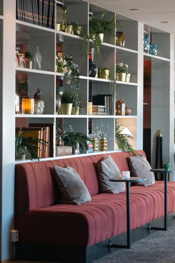 Elegant röd sammetssoffa med grå kuddar i hotellounge med bokhylla fylld av växter och dekorationer