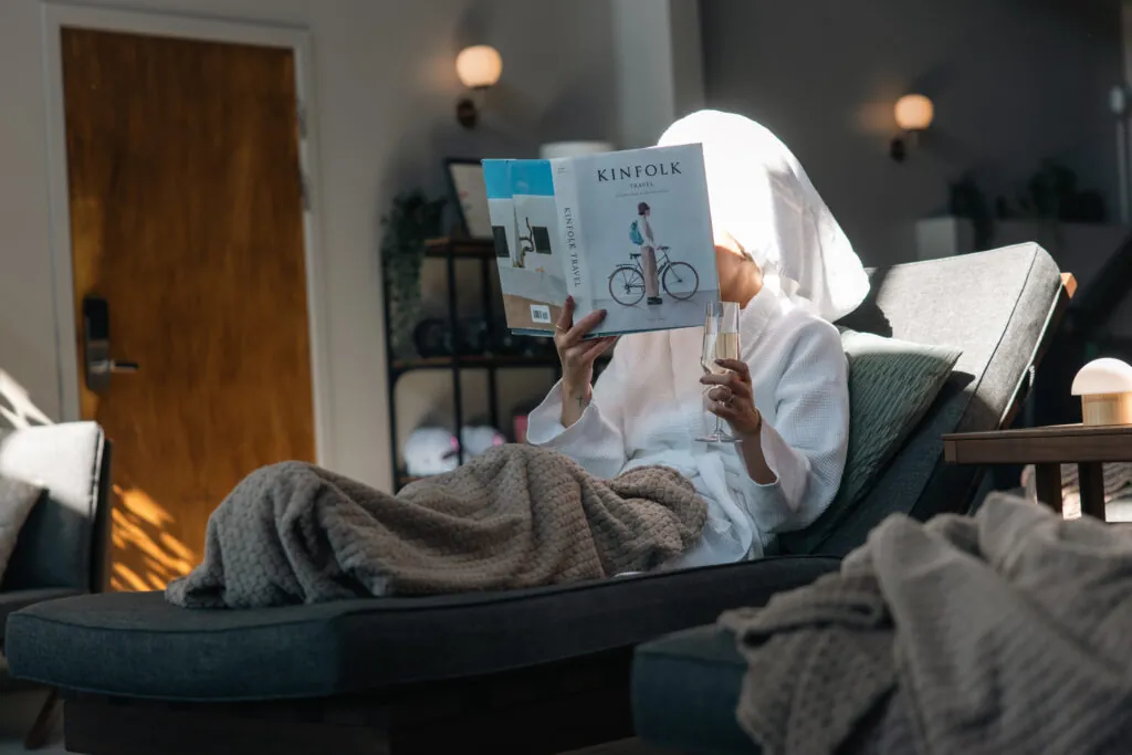 Gäst i badrock läser tidning med glas champagne i modern hotellsvit med naturgröna textilier