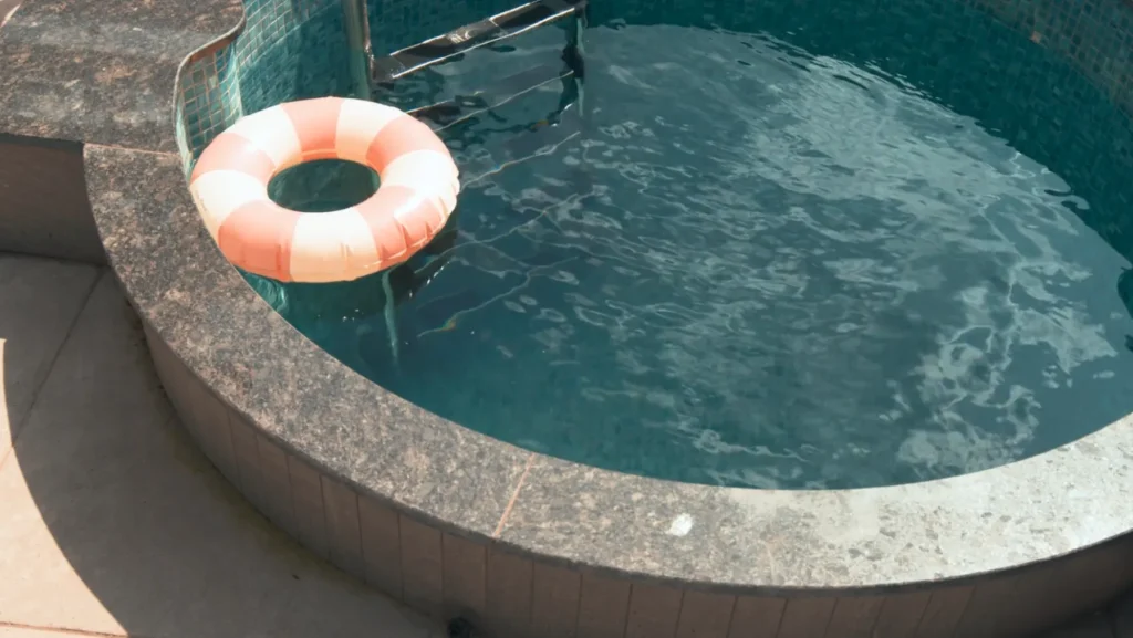 Rund bubbelpool med turkost vatten och rosa-vit uppblåsbar ring på kanten, omgiven av natursten