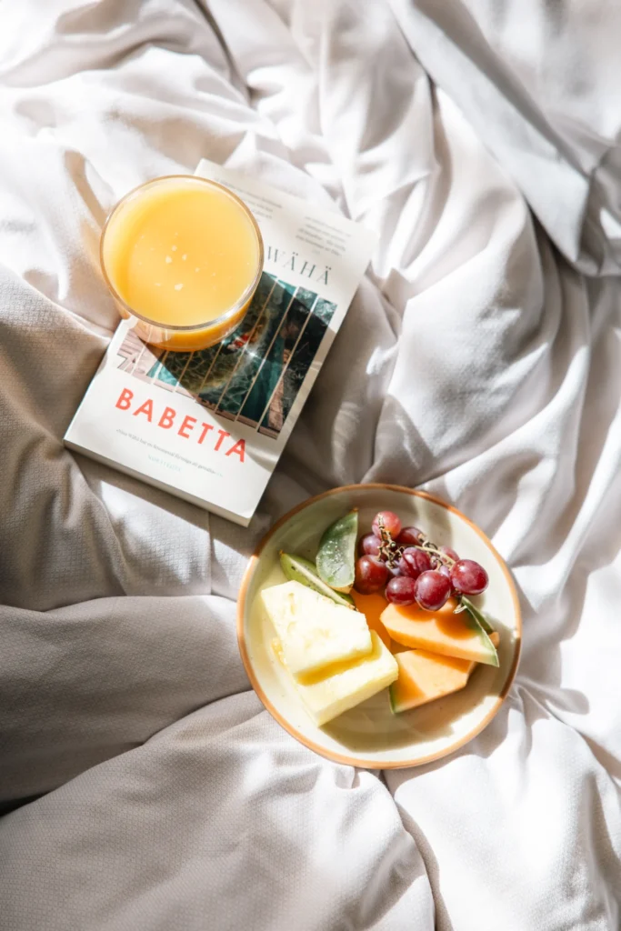 Elegantvarmt fruktfat med druvor och citrus samt färsk apelsinjuice serverat på sängen bredvid bok