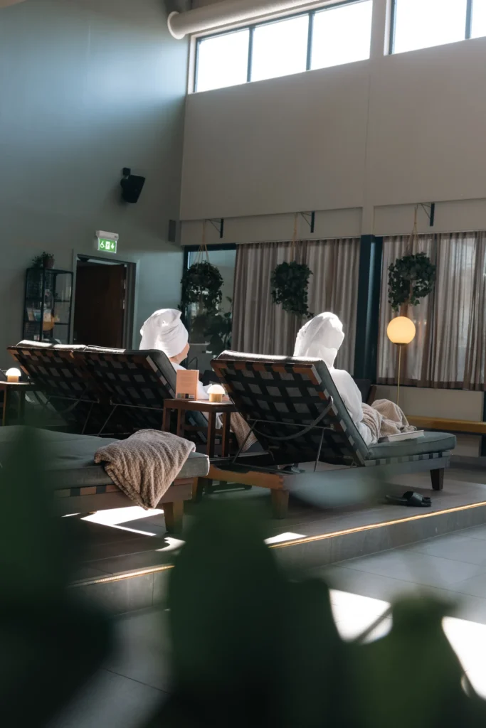 Två gäster vilar i solstolar med handdukar i spaets relaxavdelning med högt i tak och stora fönster