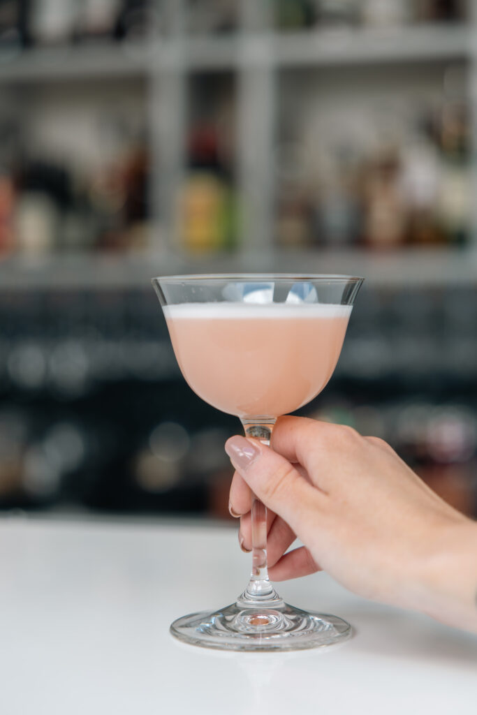 Hand hält rosa Cocktail in einem eleganten Glas vor einer Hotelbar mit Flaschen im Hintergrund