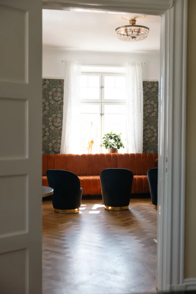 Elegant hotellsvit med orangefärgad sammetssoffa vid fönster, mörka fåtöljer och kristallkrona