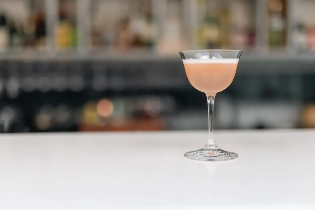 Rosa cocktail i elegant coupeglas på vit bardisk med suddig restaurangmiljö i bakgrunden