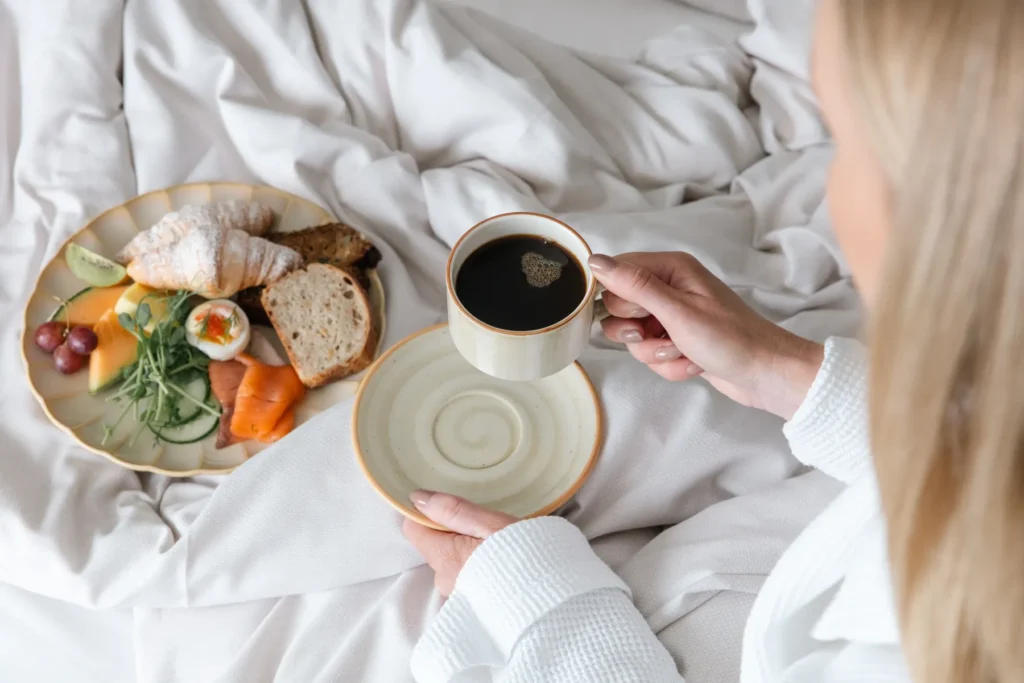 Kvinna njuter av frukost i sängen med kaffe och tallrik med croissant, ägg, frukt och grönsaker