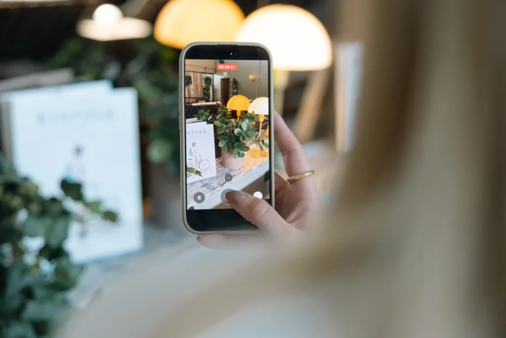Gäst fotograferar hotellrummet med mobiltelefon, visar moderna lampor och grön växtdekor i lugn miljö