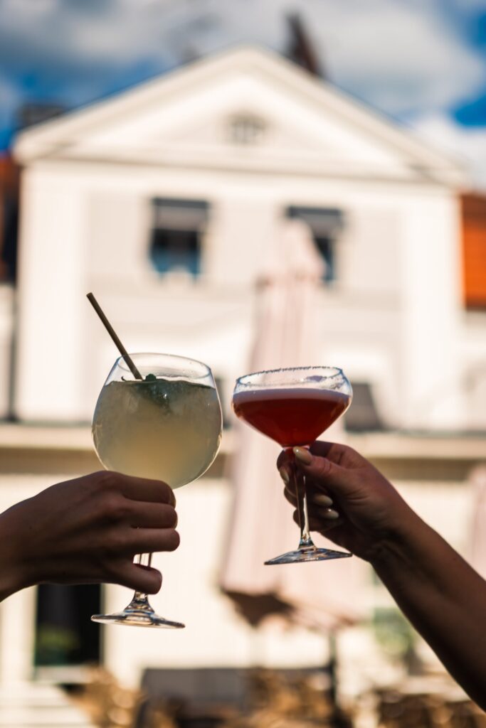 Zwei Hände stoßen mit Cocktails vor einer weißen Villa am Vätternsee an - perfekt für einen Junggesellinnenabschied mit Entspannung und Freude