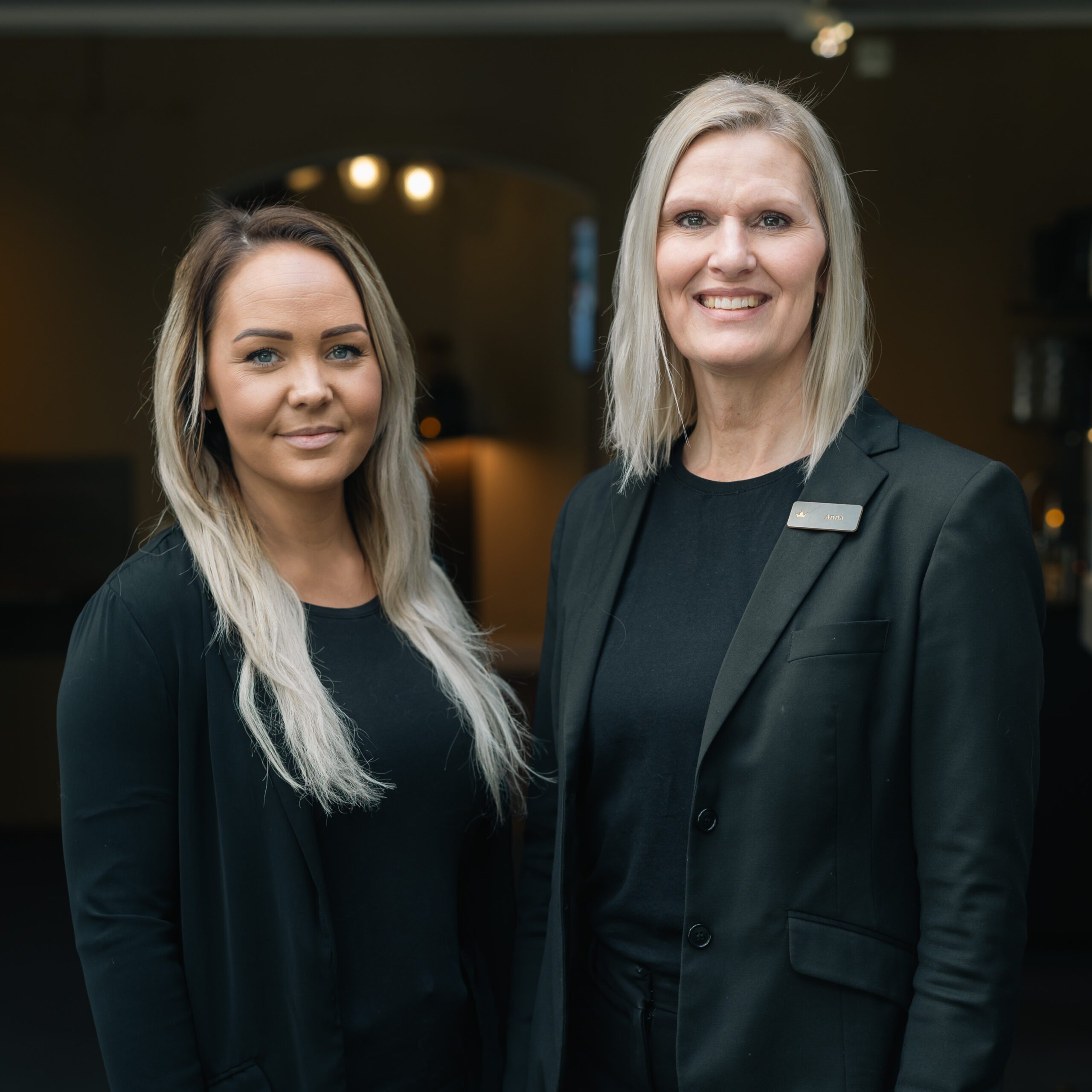 Två leende kvinnor i elegant svart klädsel står bredvid varandra, redo att välkomna möhippagäster på Starby Spa & H...