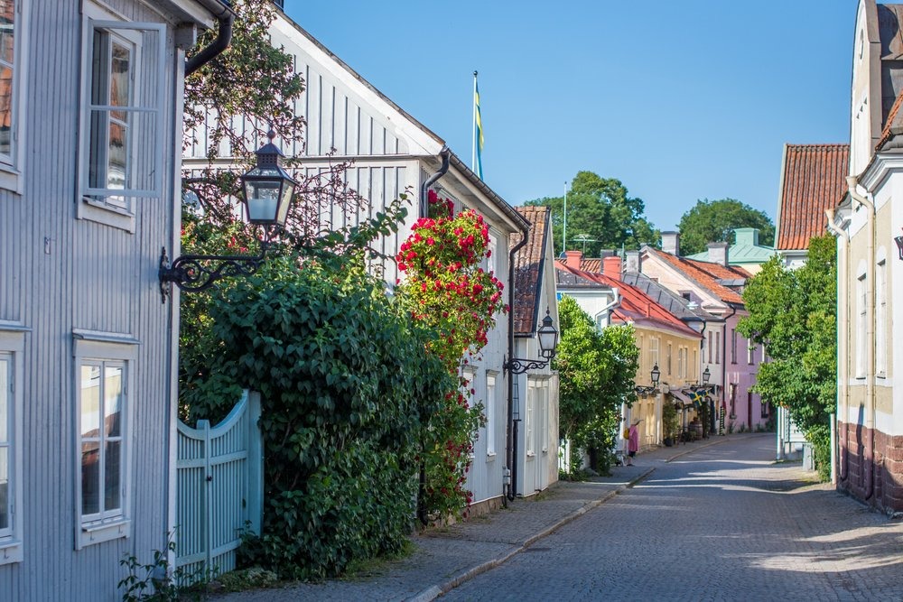 Charmig kullerstesgata i Vadstena med färgglada trähus, blomsterkorgar och svensk flagga under blå himmel