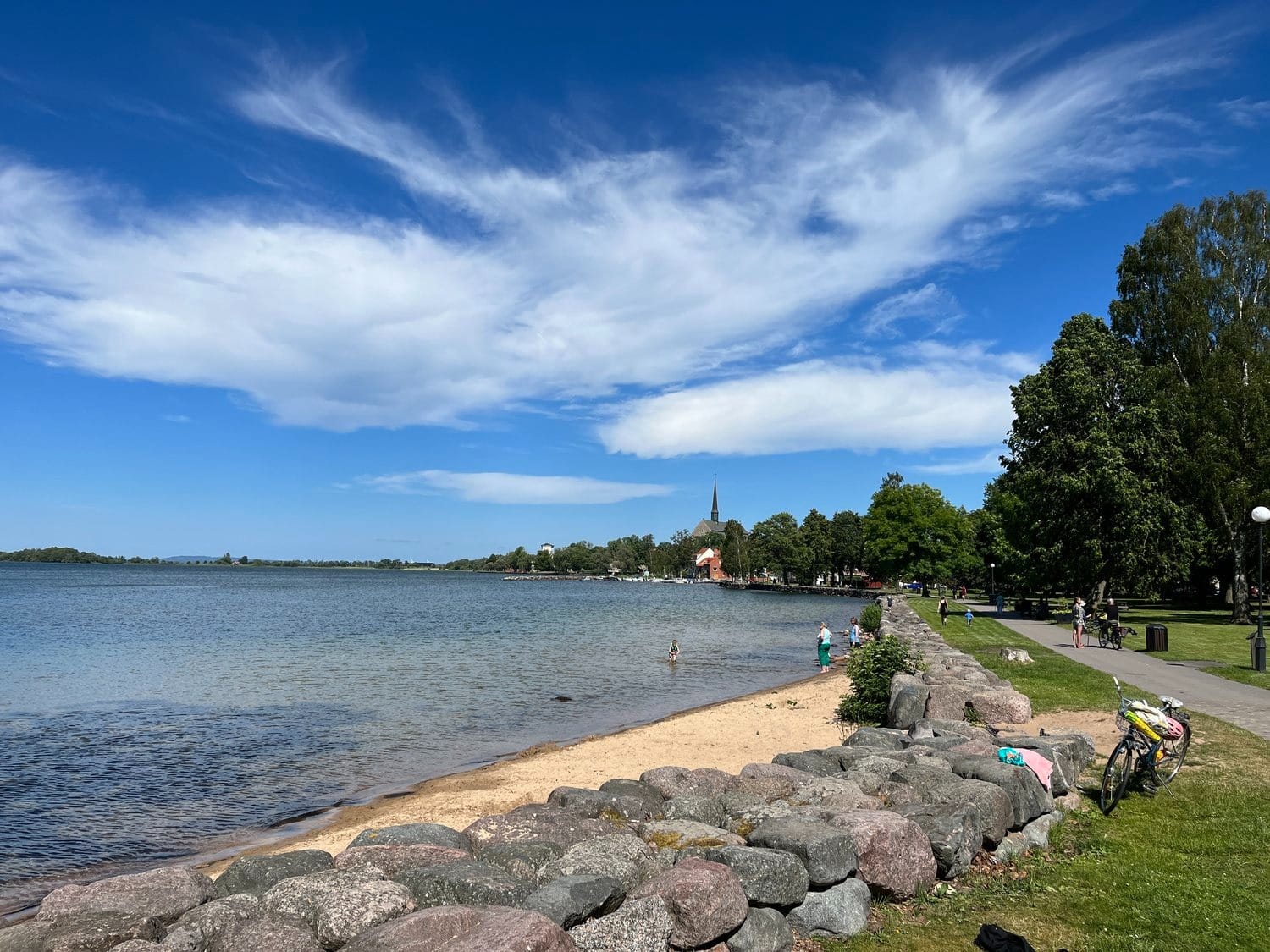 Vätterns strand vid Starby Spa & Hotell Sandstrand vid Vätterns blå vatten med stenbrygga, grön park och kyrktorn i bakgrunden under molnig himmel