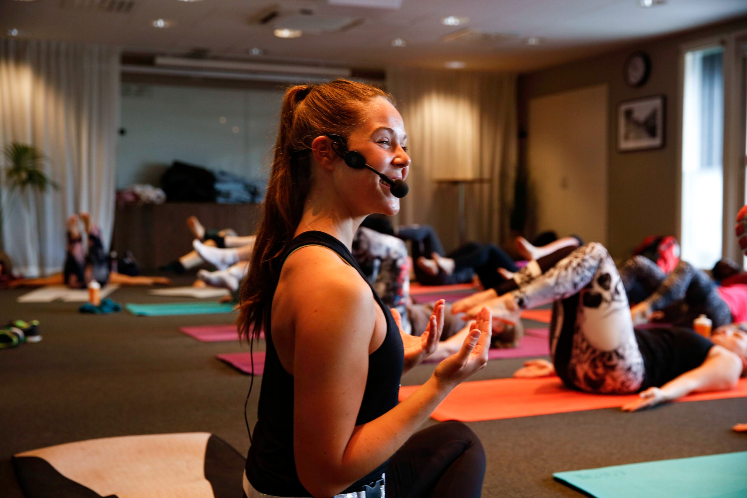 Yogainstruktör leder yogapass med mikrofon medan deltagare utövar yoga på mattor i ljus studiomiljö