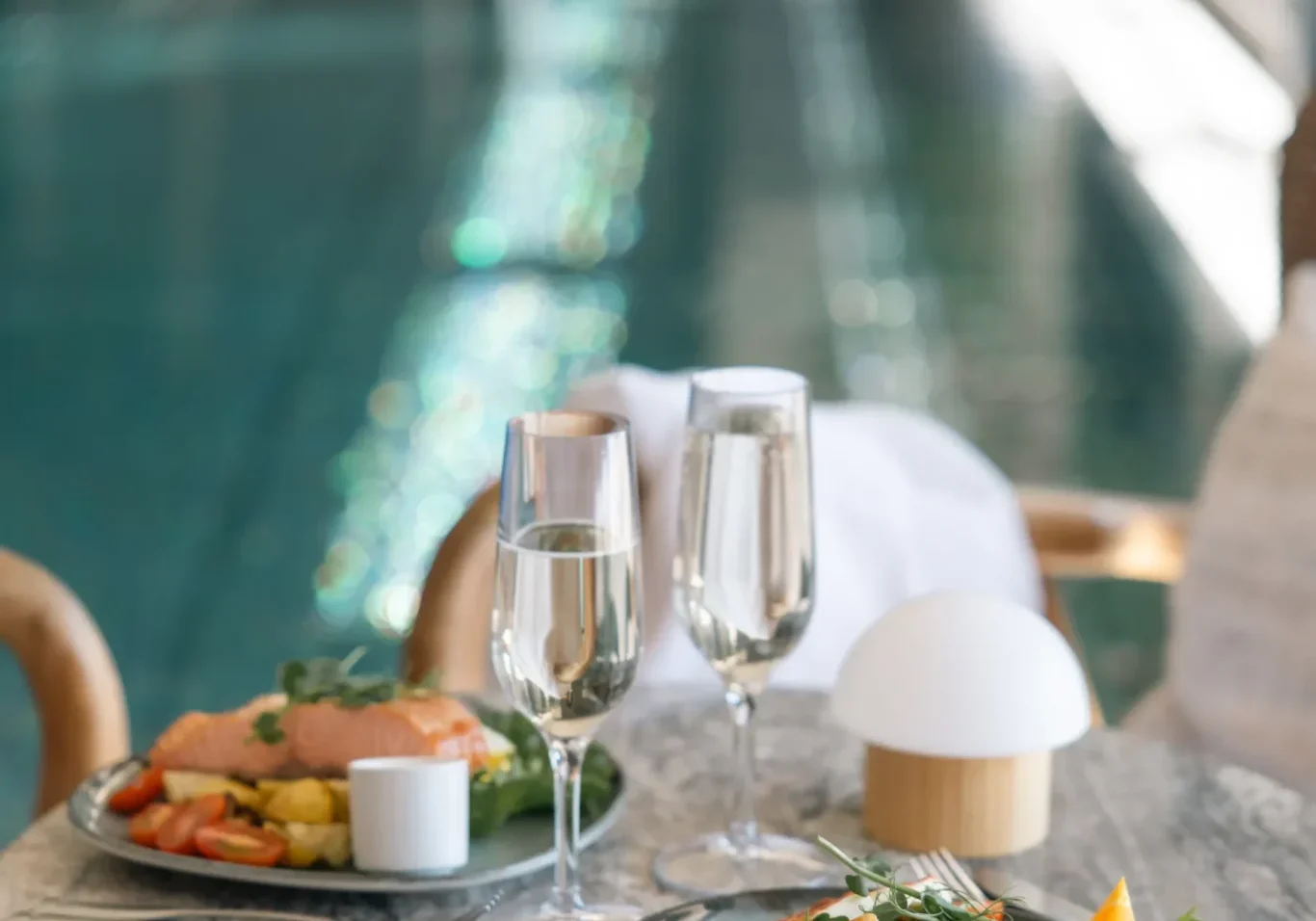 Mittags-Spa mit Essen am Pool Lunch-Spa am Innenpool im Starby Spa & Hotel – Teller mit Lachs und Halloumi, Champagnergläser und eine dezente Tischlampe