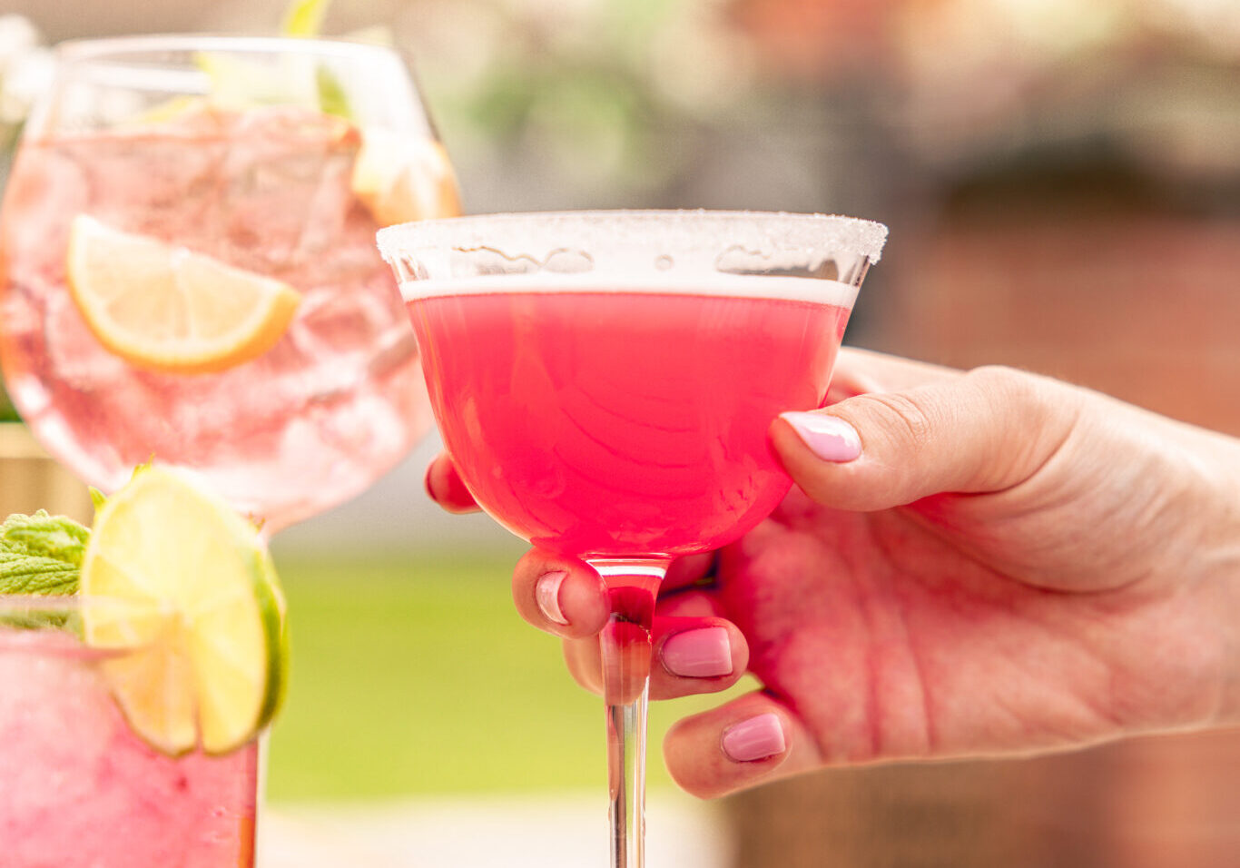 Hand håller en rosa margarita med sockrad kant vid sommardukat bord med fler cocktails och limeskivor