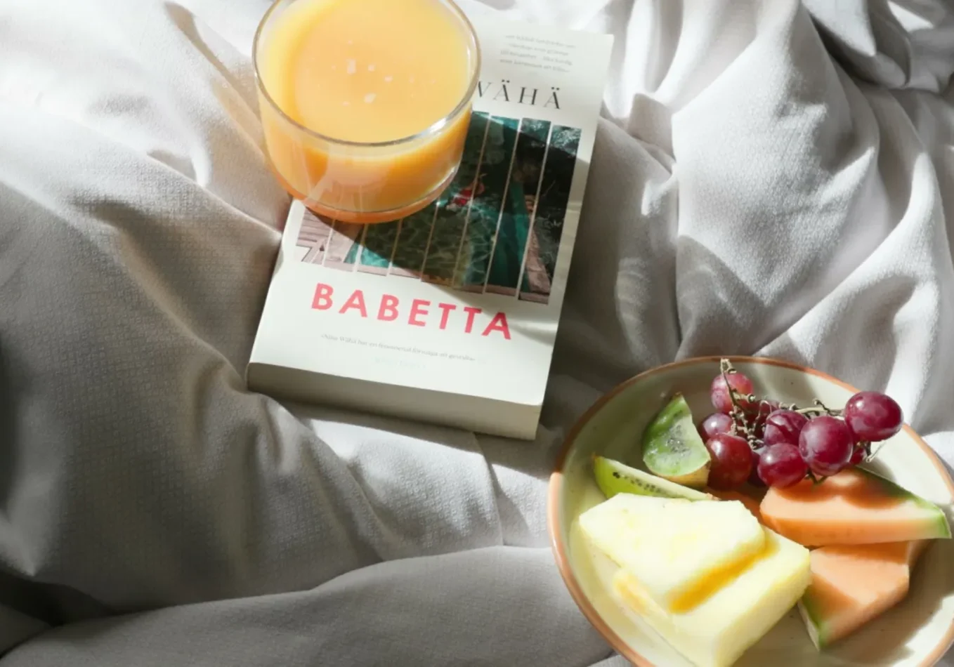 Fruktfat med ananas, melon, kiwi och druvor samt ett glas juice och boken Babetta på hotellsäng med vita lakan