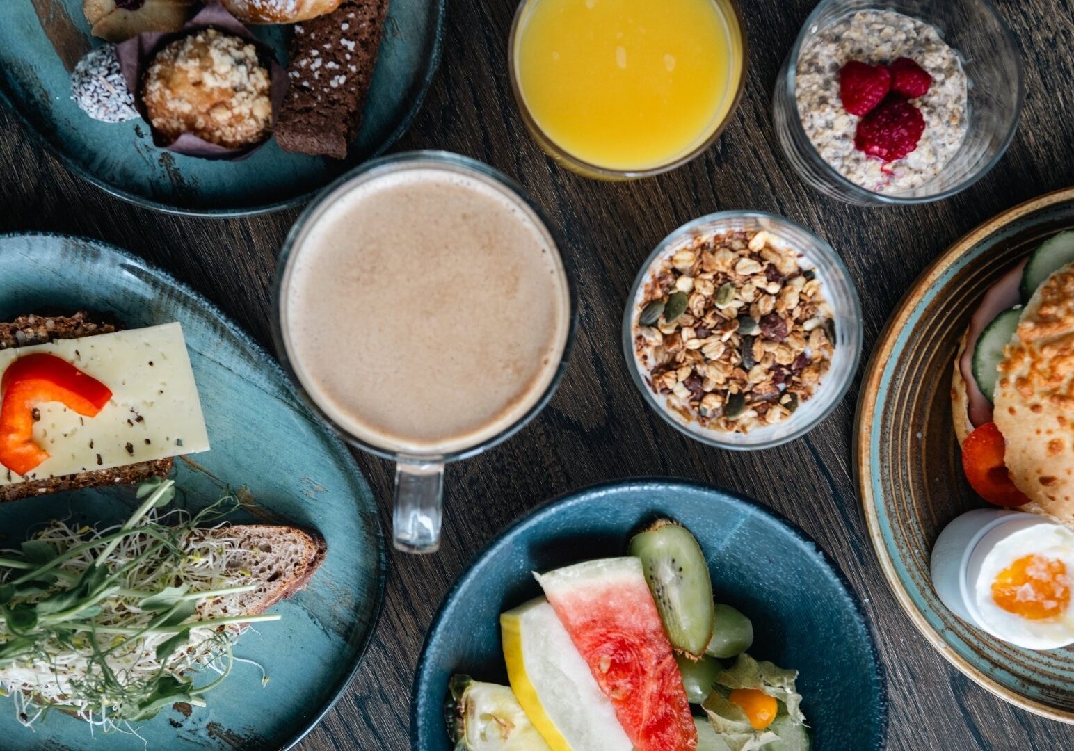 Generöst frukostbord med granola, frukt, latte, juice, ägg och smörgåsar på teal-färgade tallrikar