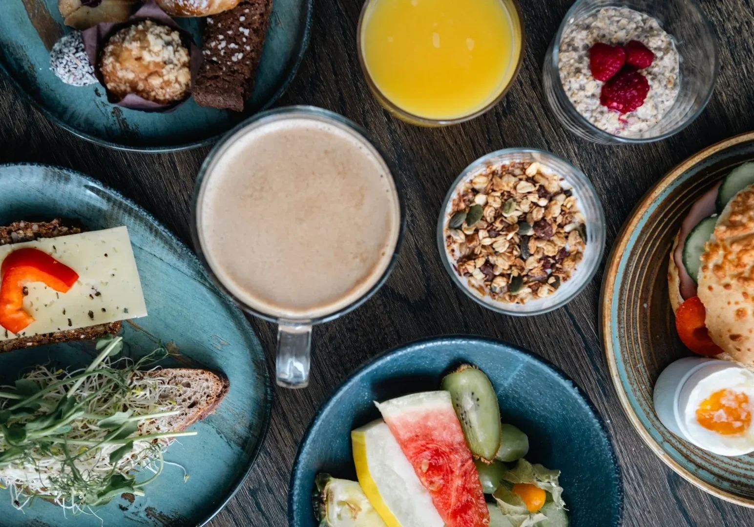 Generöst frukostbord med granola, frukt, latte, juice, ägg och smörgåsar på teal-färgade tallrikar